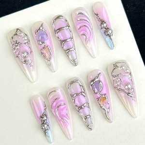 May include: A set of ten long, almond-shaped press-on nails. Each nail features a pearlescent base with a gradient of pink and purple. Silver-colored metal accents and decorative stones adorn each nail, creating a unique and artistic design.