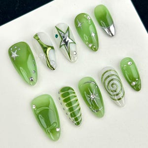以下が含まれることがあります： 緑と白の人工爪10本セット。星のアクセント、シルバーの先端、スパイラル模様など、さまざまなデザインが施されています。一部の爪にはラインストーンが埋め込まれています。爪は楕円形です。
