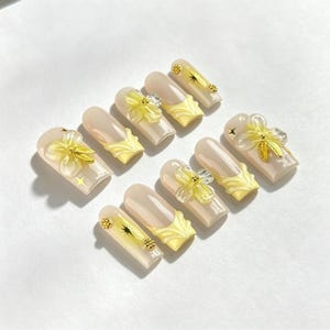 May include: A set of ten press-on nails featuring a nude base with yellow French tips. The design includes three-dimensional translucent yellow flower embellishments, small gold metallic star accents, and tiny gold circular studs on the nail surface.