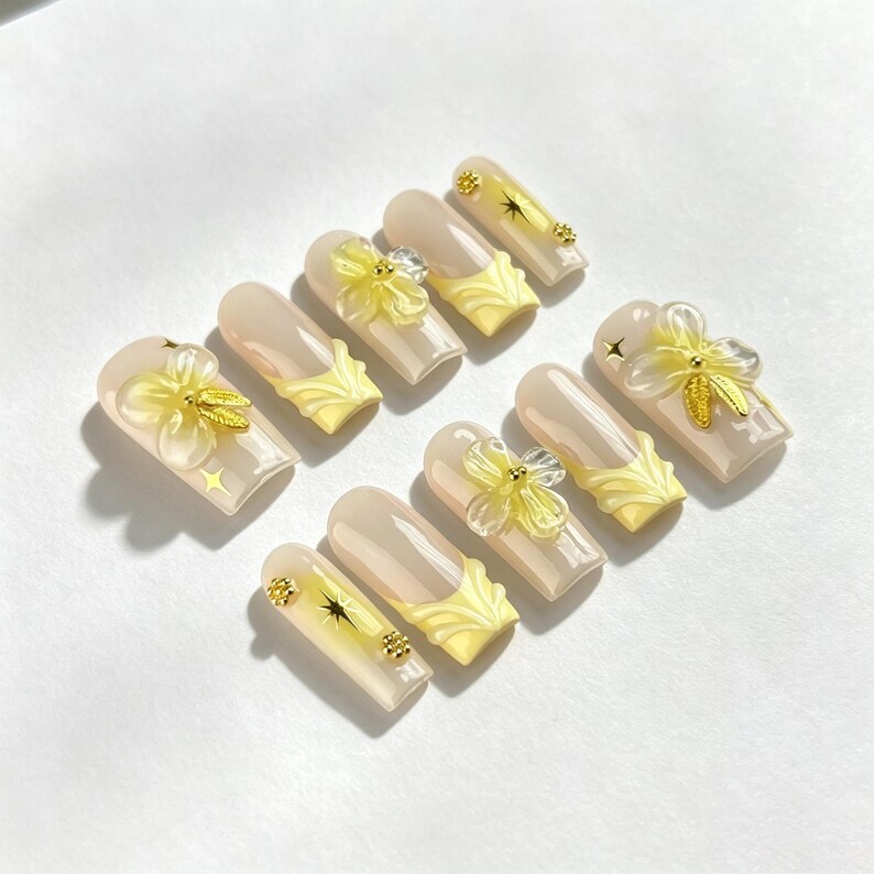 May include: A set of ten press-on nails featuring a nude base with yellow French tips. The design includes three-dimensional translucent yellow flower embellishments, small gold metallic star accents, and tiny gold circular studs on the nail surface.