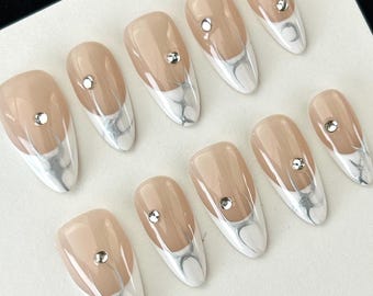 May include: Set of artificial nails in a neutral beige color with a white and silver marble design at the tips. Each nail features a small, round, silver gem. The nails are oval-shaped.