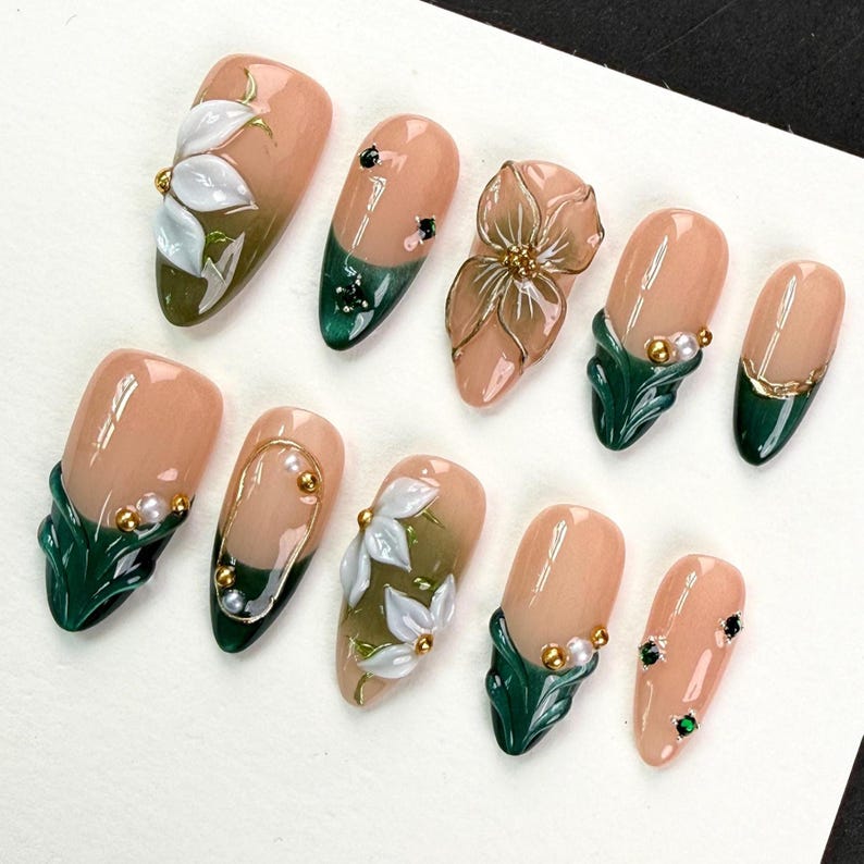 May include: A set of ten press-on nails featuring a nude base with dark green tips. The designs include three-dimensional white floral accents, gold metallic lines, small white pearls, and tiny green and gold studs arranged in various patterns.