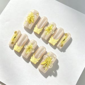 May include: A set of ten press-on nails featuring a glossy nude base with pale yellow French tips. The design includes three-dimensional translucent yellow flowers, gold metallic leaf accents, and small gold star and dot embellishments.