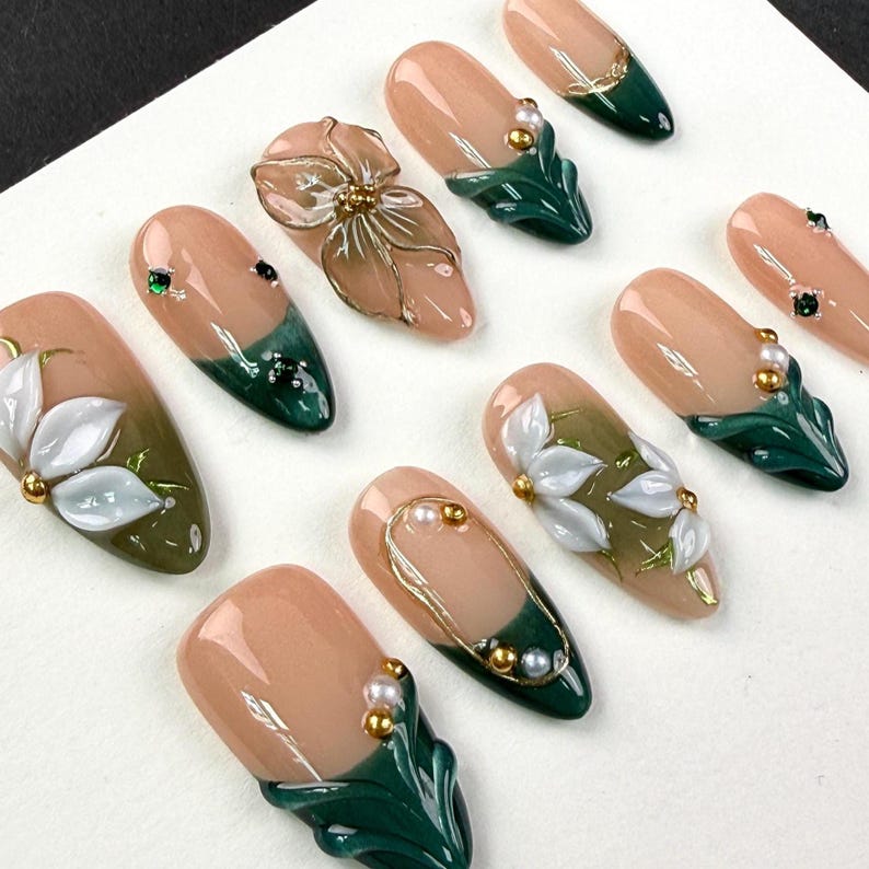 May include: A set of ten press-on nails featuring a nude base with dark green tips. The designs include three-dimensional white floral accents, gold metallic detailing, and small embedded pearls and rhinestones on an almond-shaped nail surface.