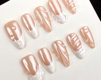 May include: A set of ten artificial nails in various designs. The nails feature a pearlescent peach color with white accents. Some nails have a French tip design, while others have textured patterns or small, raised embellishments.