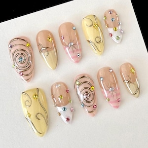 May include: A collection of ten artificial nails with diverse designs. The nails showcase light pink, yellow, and nude hues, embellished with silver swirls, star-shaped details, and colourful rhinestones. The nails are oval-shaped.