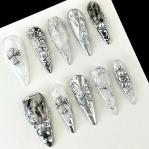 Könnte beinhalten: Eine Sammlung von zehn langen, spitzen künstlichen Nägeln. Die Nägel weisen eine Vielzahl von Designs auf, darunter silberne Kreuze, Schmetterlingsakzente und Perlenverzierungen. Einige Nägel haben einen Marmoreffekt, andere sind klar.