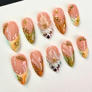 May include: A set of ten press-on nails featuring a nude base with olive green and orange accents. The design includes raised 3D flower embellishments, small gold metallic spheres, and gold star-shaped charms arranged across the nail surfaces.