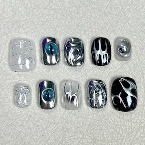 May include: A set of ten artificial nails in various designs. The nails feature silver, black, and white colors with unique patterns, including an eye design, abstract swirls, and cracked textures. The nails are oval-shaped and have a glossy finish.