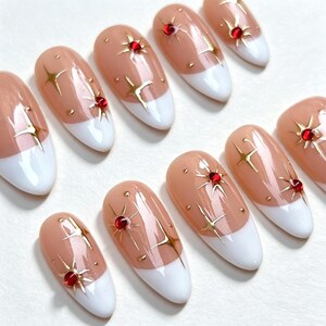 May include: A set of almond-shaped press-on nails featuring a nude base with white French tips. Each nail is decorated with gold starburst accents and small, round, deep red rhinestones placed at the center of some of the star designs.