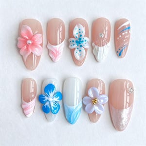 May include: A set of ten press-on nails featuring a nude base with various designs including 3D pink and blue floral accents, white and blue swirls, painted dots, and small clear rhinestones. The nails have an almond shape and glossy finish.
