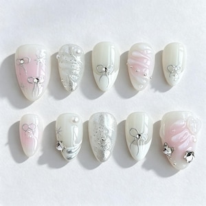 May include: A set of ten press-on nails featuring a white and soft pink color palette. The designs include silver metallic bow accents, raised 3D swirls, silver glitter, small white pearls, and clear crystal embellishments on various nail tips.