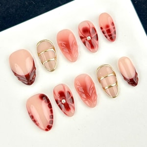 May include: A set of ten artificial nails in various designs. The nails feature a gradient of pink and red hues, with some adorned with gold accents or intricate patterns. Some nails have a jewel embedded in the design.