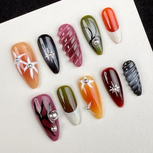 May include: A collection of ten artificial nails in various designs and colors. The nails feature a range of styles, including solid colors, intricate patterns, and embellishments like silver studs. Colors include orange, black, green, and red.