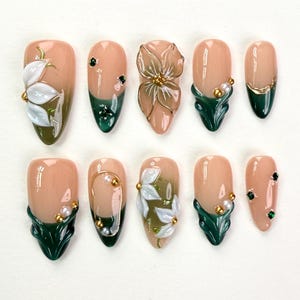 May include: A set of ten press-on nails featuring a nude base with dark green tips. The designs include three-dimensional white floral accents, small gold beads, white pearls, and tiny dark green rhinestones arranged in various patterns.