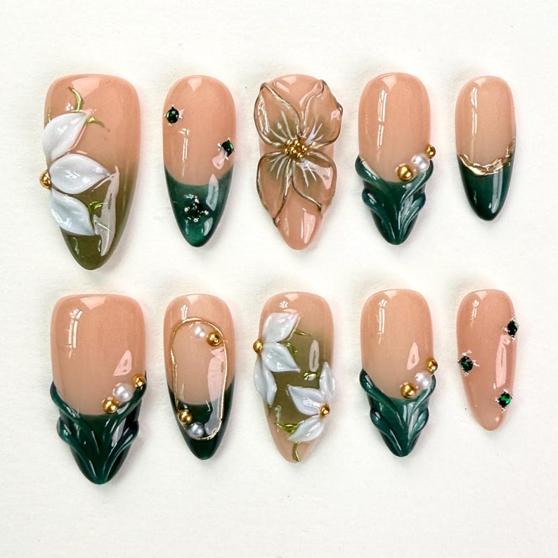 May include: A set of ten press-on nails featuring a nude base with dark green tips. The designs include three-dimensional white floral accents, small gold beads, white pearls, and tiny dark green rhinestones arranged in various patterns.