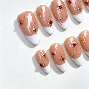May include: A set of almond-shaped press-on nails featuring a nude base with white French tips. Each nail is decorated with gold starburst accents, small gold dots, and tiny red rhinestones centered within some of the starburst designs.