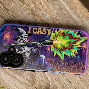 May include: A rectangular phone case featuring a graphic of a wizard in a pointed hat firing a rocket launcher. The design includes a vibrant green, yellow, and red explosion effect and the text I CAST NON-MAGIC MISSILE in bold yellow letters.