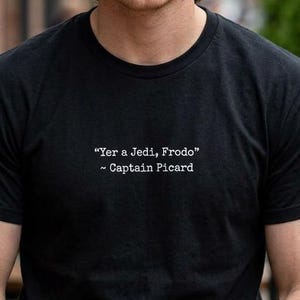 May include: Black crew neck t-shirt with the quote "Yer a Jedi, Frodo" attributed to Captain Picard in white text. The person is sitting at a wooden table outside.