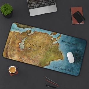 Peut inclure: Tapis de souris rectangulaire avec une carte détaillée de Ferelden, avec le texte "Ferelden" en évidence. La carte est dans des tons de marron, de vert et de bleu, avec des caractéristiques géographiques étiquetées. Une souris blanche et d'autres fournitures de bureau sont visibles.