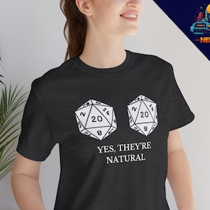 Puede incluir: Una camiseta negra con un gráfico blanco de dos dados de veinte caras. El texto "YES, THEY'RE NATURAL" está impreso debajo de los dados.