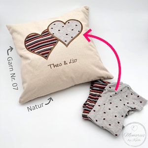 Könnte beinhalten: Ein beiges Kissen mit zwei gestickten Herzen, eines mit einem gestreiften Muster und das andere mit einem gepunkteten Muster. Das Kissen trägt den gestickten Text "Theo & Liv". Das Kissen liegt neben zwei Stoffresten, einer mit einem gestreiften Muster und der andere mit einem gepunkteten Muster.