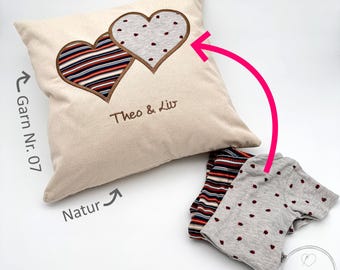 May include: A beige pillow with two embroidered hearts, one with a striped pattern and the other with a dotted pattern. The pillow has the text "Theo & Liv" embroidered on it. The pillow is next to two fabric scraps, one with a striped pattern and the other with a dotted pattern.