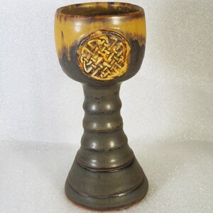 May include: A handcrafted ceramic chalice with a Celtic knot design. The chalice features a two-tone glaze, with a yellow-brown rim and a dark green body. The stem has a ribbed design, and the base is flared.