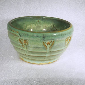 May include: A handmade ceramic bowl with a green glaze and embossed Celtic knot designs. The bowl has a rounded shape with a slightly flared rim and horizontal ridges. The interior has a lighter green hue.