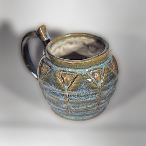 May include: A handmade ceramic mug with a unique design. The mug features a dark blue and brown glaze with a textured surface and geometric patterns. The handle is also glazed in a dark blue color. This mug is perfect for coffee or tea.