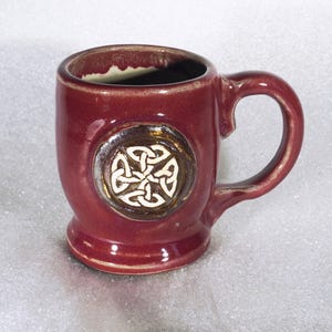 May include: A burgundy ceramic mug with a Celtic knot design. The mug has a rounded body, a handle, and a decorative emblem with a white knot design on a brown background. The mug is suitable for hot beverages.