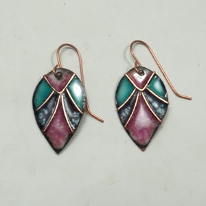 May include: A pair of leaf-shaped earrings with copper hooks. Each earring features a teal and magenta enamel design with black and copper accents. The earrings have a handmade, artisan appearance.