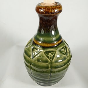 May include: Green ceramic soap dispenser with a cream-colored pump. The dispenser has a bulbous base with embossed Celtic knot designs and a dark brown neck. The top is brown and the pump is cream-colored.