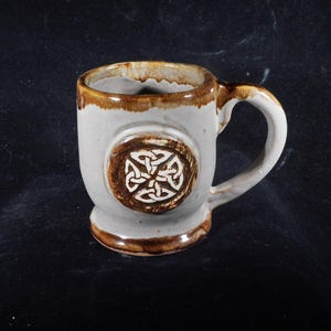May include: A ceramic mug with a Celtic knot design. The mug is primarily gray with brown accents on the rim, base, and handle. A circular emblem with a white Celtic knot is centered on the mug's body.