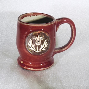 May include: A burgundy ceramic mug with a handle and a decorative thistle emblem. The mug has a wide base and a slightly flared rim. The thistle emblem is white and set in a circular, textured frame.