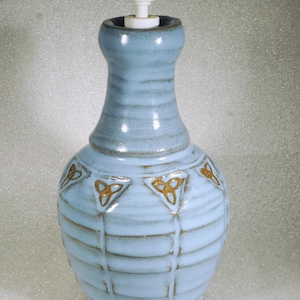 May include: A light blue ceramic soap dispenser with a white pump. The dispenser has a bulbous base with a series of horizontal ridges and gold-colored Celtic knot designs. The top of the dispenser is a narrower, cylindrical shape.