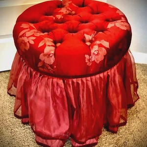 May include: A round, red ottoman with a tufted top featuring a floral pattern. The base is draped with a ruffled, sheer red fabric. The ottoman is a decorative furniture piece.