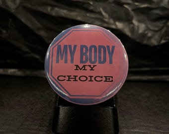 Può includere: Un bottone rosa e blu con il testo "MY BODY MY CHOICE" in lettere nere.