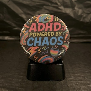 Może przedstawiać: Okrągły przycisk z czarnym tłem i kolorowym wzorem. Przycisk głosi "ADHD: Powered by Chaos!" z tęczą, błyskawicami i filiżanką kawy.