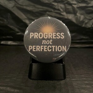 May include: A round button with a black background and a white sunburst design. The text "PROGRESS not PERFECTION" is printed in white on the button.