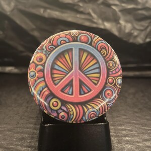 Könnte beinhalten: Ein bunter Peace-Zeichen-Button mit psychedelischem Design. Der Button zeigt ein pinkfarbenes Peace-Zeichen mit blauem Rand und einem regenbogenfarbenen Hintergrund. Das Peace-Zeichen ist von einem wirbelnden Muster aus Gelb, Blau und Pink umgeben.