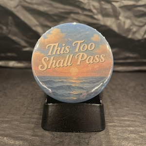 May include: A blue and orange button with a sunset scene and the text "This Too Shall Pass".