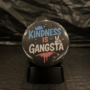 May include: A circular pin-back button with a black background featuring the text "KINDNESS IS GANGSTA" in white and light blue. The design includes a light blue paint splatter, a pink paint drip, a peace sign hand gesture, and small orange stars.