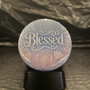 May include: A round button with a blue and purple ombre background. The button has a white floral design and the word "Blessed" in white cursive lettering.