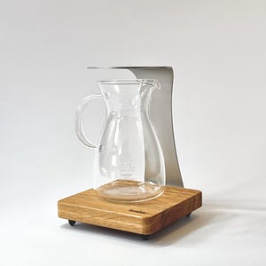 Könnte beinhalten: Eine klare Glaskaraffe mit Griff und Ausguss steht auf einem Holzsockel mit Metallhalterung. Die Karaffe ist zum Brühen von Kaffee oder Tee bestimmt. Der Holzsockel hat eine natürliche Holzmaserung.