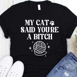 May include: Black t-shirt with the text "MY CAT SAID YOU'RE A BITCH" in white, along with a paw print and yarn ball graphic. The shirt is paired with denim shorts and white sneakers, set against a white wooden background.