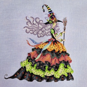 Puede incluir: Diseño de punto de cruz de una bruja con pelo largo y gris y un sombrero a rayas adornado con flores. Viste un vestido en capas en naranja, verde y negro con lunares y sostiene una escoba. El fondo es azul claro.