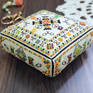 Pode incluir: Uma caixa quadrada de ponto de agulha esbranquiçada com um design detalhado de ponto cruz. A caixa apresenta um padrão floral central e bordas com tesouras, bobinas e outros motivos relacionados à costura em laranja, verde, roxo e preto. As bordas são adornadas com pequenas contas coloridas.