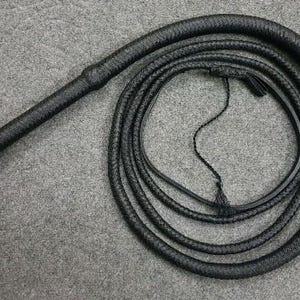 Puede incluir: Un látigo de cuero negro con un mango trenzado. El látigo está enrollado y listo para usar.