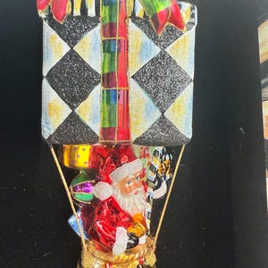 Puede incluir: Adorno navideño con forma de globo aerostático con Papá Noel en la cesta. El globo es una caja de regalo con un patrón de cuadros blancos y negros. El adorno tiene detalles rojos, verdes y dorados.